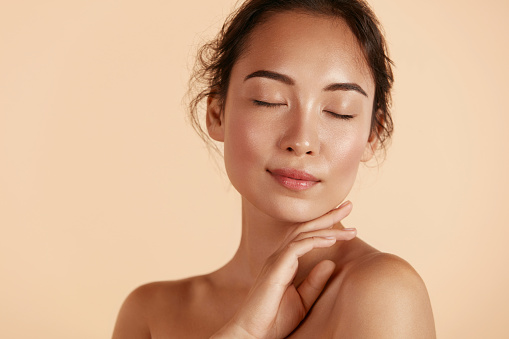 Beauty face. Woman with natural makeup and healthy skin portrait. Beautiful asian girl model touching fresh glowing hydrated facial skin on beige background closeup. Skin care concept